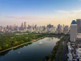 An aerial skyline view of Benjakitti Park lake in Bangkok against a hazy sunrise sky.